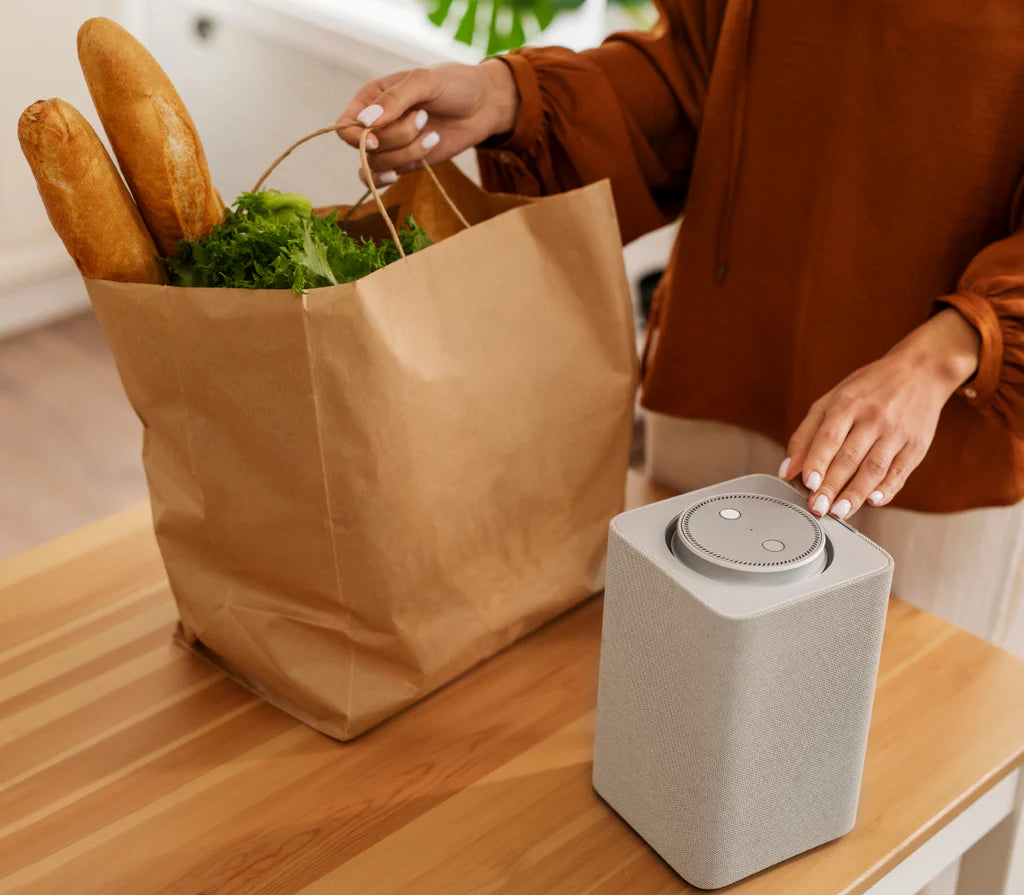 Person with a brown paper bag of groceries next to a smart speaker on a wooden table.